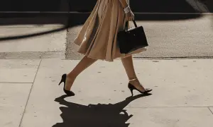 Elegancka kobieta w beżowej sukience i czarnych szpilkach, niosąca torebkę. Idealne buty do beżowej sukienki.