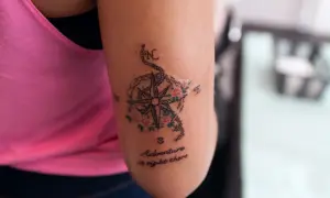 Tatuaż róża wiatrów z kwiatami i napisem "Adventure is right there" na ramieniu.
