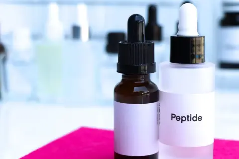 Dwie buteleczki z pipetkami, jedna z napisem "Peptide". W tle inne kosmetyki.