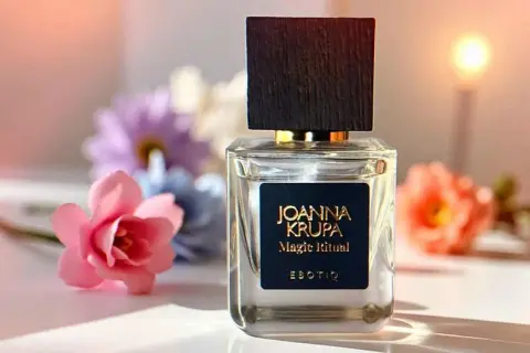 Elegancka butelka perfum Joanna Krupa Magic Ritual na tle kwiatów. Wiele osób szuka magiczne perfumy opinie, by poznać ten zapach.