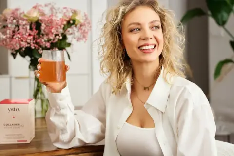 Uśmiechnięta kobieta z kręconymi blond włosami trzyma szklankę napoju. Obok stoi pudełko z napisem "Collagen Your Skin", sugerujące kolagen hydrolizowany.