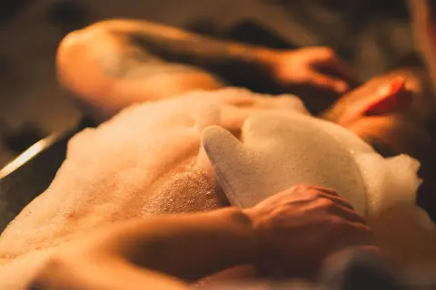 Ciepła piana otula ciało podczas rytuału hammam. Ręka z rękawicą do masażu spoczywa na skórze, przygotowując do głębokiego relaksu.