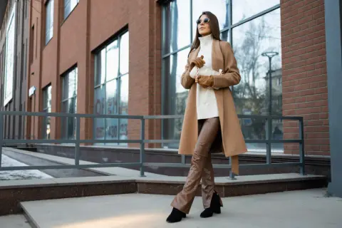 Elegancka kobieta w płaszczu w kolorze camel i brązowych spodniach ze skóry ekologicznej pozuje na tle nowoczesnego budynku.