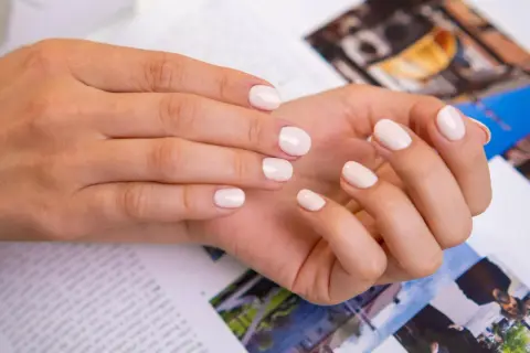 Delikatne, modne paznokcie french w odcieniu nude. Dłonie spoczywają na magazynie z kolorowymi zdjęciami.