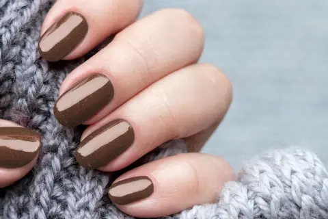 Dłonie z perfekcyjnym manicurem kombinowanym w odcieniu czekolady, otulone miękkim, szarym swetrem.
