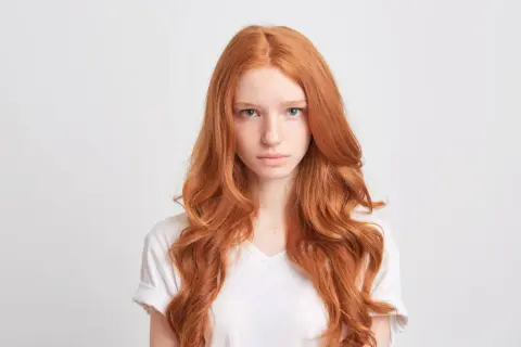Dziewczyna z burzą włosów w odcieniu rudy, z piegami na nosie, patrzy prosto przed siebie.