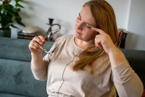 Kobieta z okładami na oczy, masuje skroń, trzymając okulary.