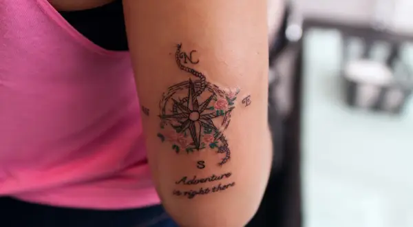 Tatuaż róża wiatrów z kwiatami i napisem "Adventure is right there" na ramieniu.
