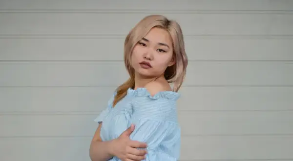 Młoda kobieta z blond włosami w niebieskiej sukience w paski, na tle białej ściany.