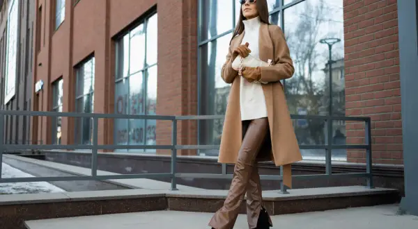 Elegancka kobieta w płaszczu w kolorze camel i brązowych spodniach ze skóry ekologicznej pozuje na tle nowoczesnego budynku.
