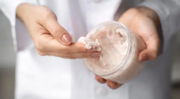 Dłonie w białym kitlu pobierają palcem drobnoziarnisty peeling. W słoiczku widać jego bogatą konsystencję, która może zawierać nawilżającą **urea**.