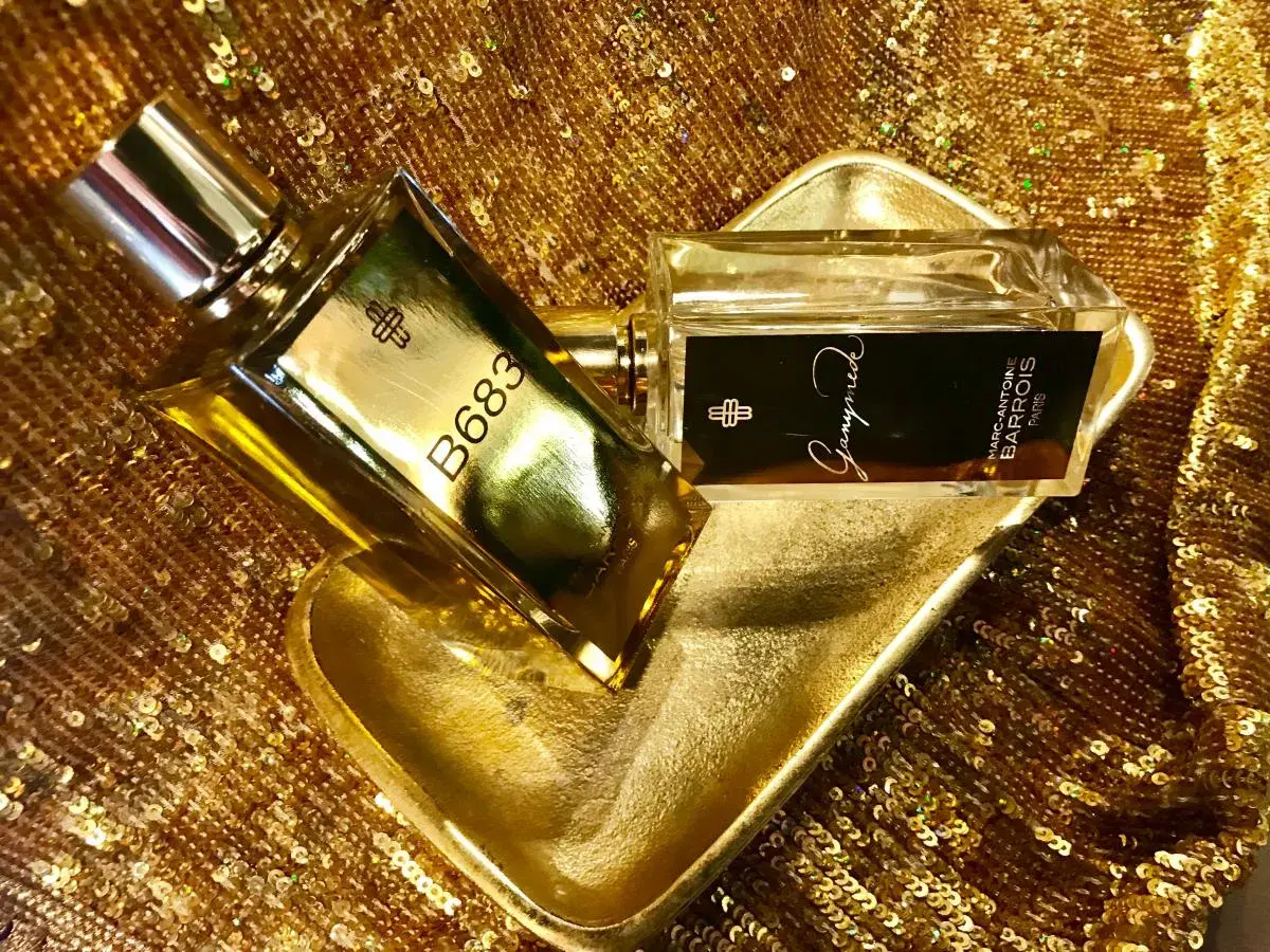 Dwa flakony perfum Marc Antoine Barrois na złotej, cekinowej tkaninie. Jeden flakon z napisem B683, drugi z nazwą "Ganymede".