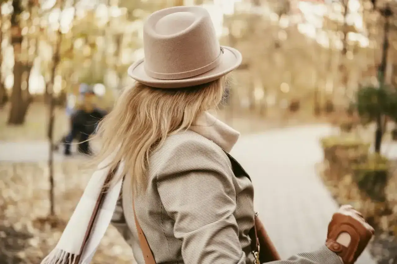Elegancki toczek na głowie blondynki, w jesiennym parku. Wiatr rozwiewa jej włosy, a ona w rękawiczkach idzie ścieżką.