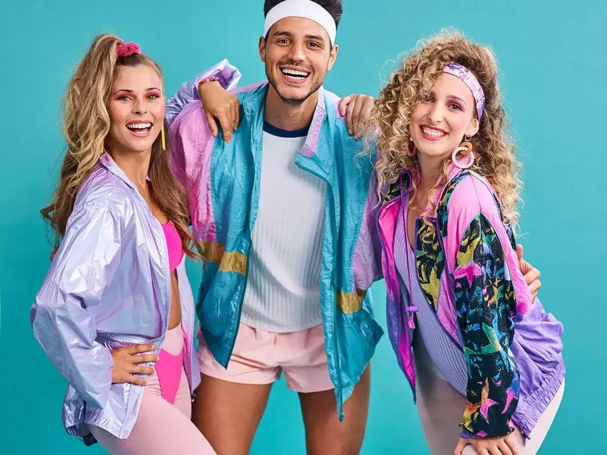 Trzy uśmiechnięte osoby w kolorowych strojach sportowych, żywcem wyjęte z lat 80. Ich styl to kwintesencja mody tamtej dekady.