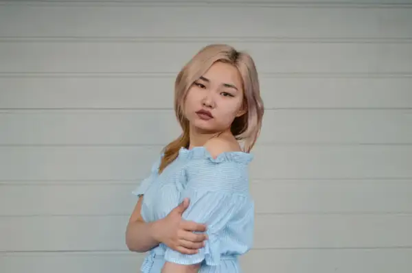 Młoda kobieta z blond włosami w niebieskiej sukience w paski, na tle białej ściany.