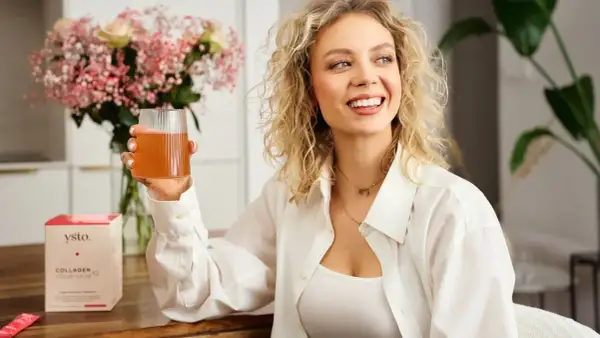 Uśmiechnięta kobieta z kręconymi blond włosami trzyma szklankę napoju. Obok stoi pudełko z napisem "Collagen Your Skin", sugerujące kolagen hydrolizowany.