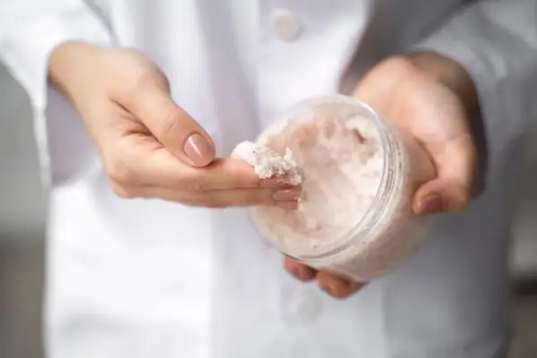 Dłonie w białym kitlu pobierają palcem drobnoziarnisty peeling. W słoiczku widać jego bogatą konsystencję, która może zawierać nawilżającą **urea**.